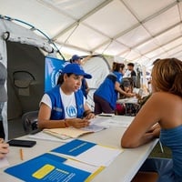 Detrás de una mesa, una trabajadora de ACNUR con vestimenta azul de la organización, atiende a una mujer que está de espaldas del otro lado de la mesa