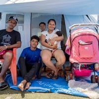 Una familia de cinco miembros, entre ellos un adolescente, un niño y una bebé, está sentada frente a la entrada de una tienda de campaña
