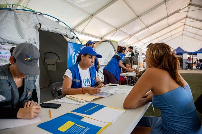 Detrás de una mesa, una trabajadora de ACNUR con vestimenta azul de la organización, atiende a una mujer que está de espaldas del otro lado de la mesa