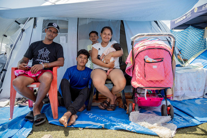 Una familia de cinco miembros, entre ellos un adolescente, un niño y una bebé, está sentada frente a la entrada de una tienda de campaña