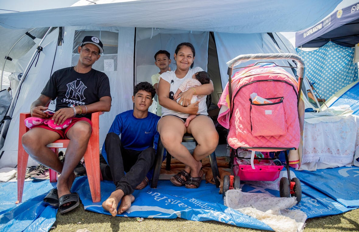 Una familia de cinco miembros, entre ellos un adolescente, un niño y una bebé, está sentada frente a la entrada de una tienda de campaña