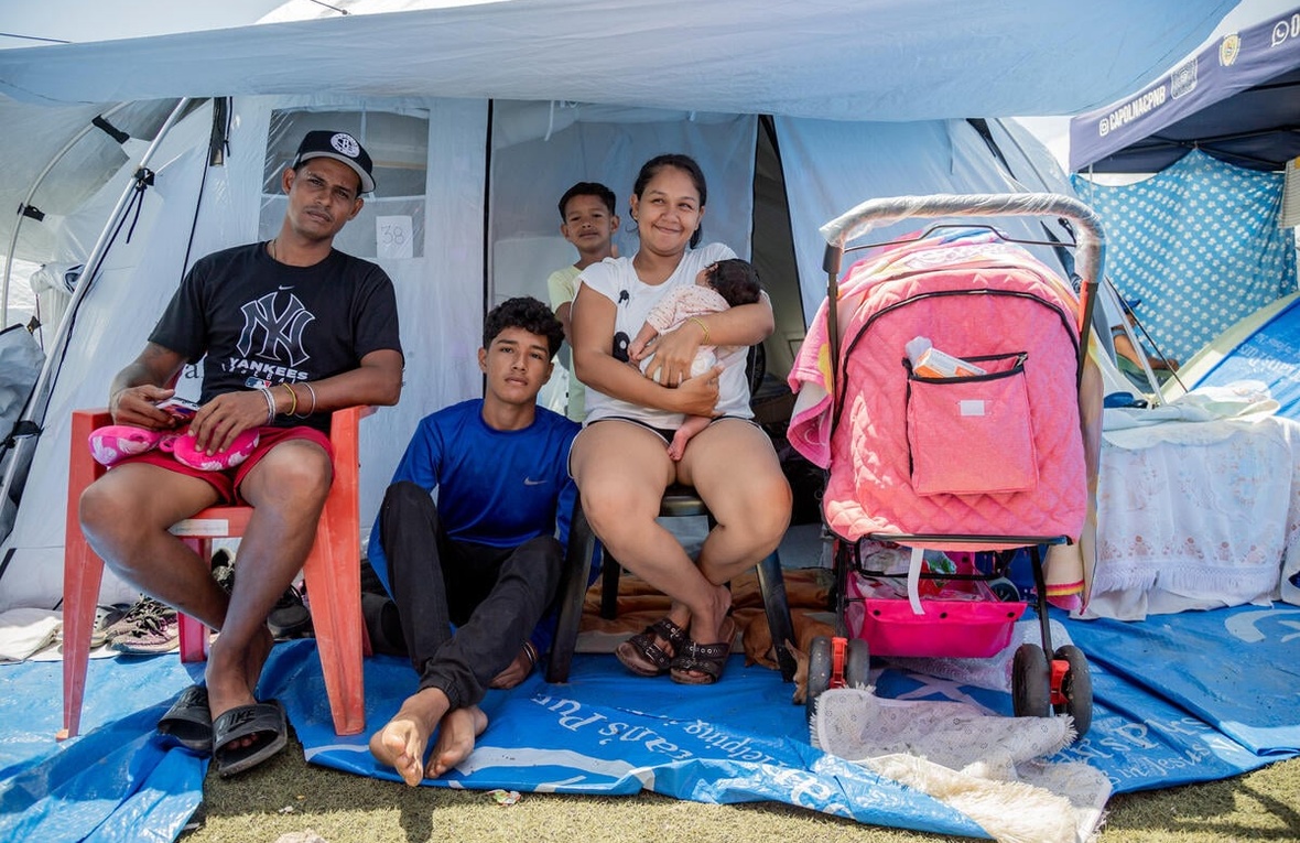 Una familia de cinco miembros, entre ellos un adolescente, un niño y una bebé, está sentada frente a la entrada de una tienda de campaña
