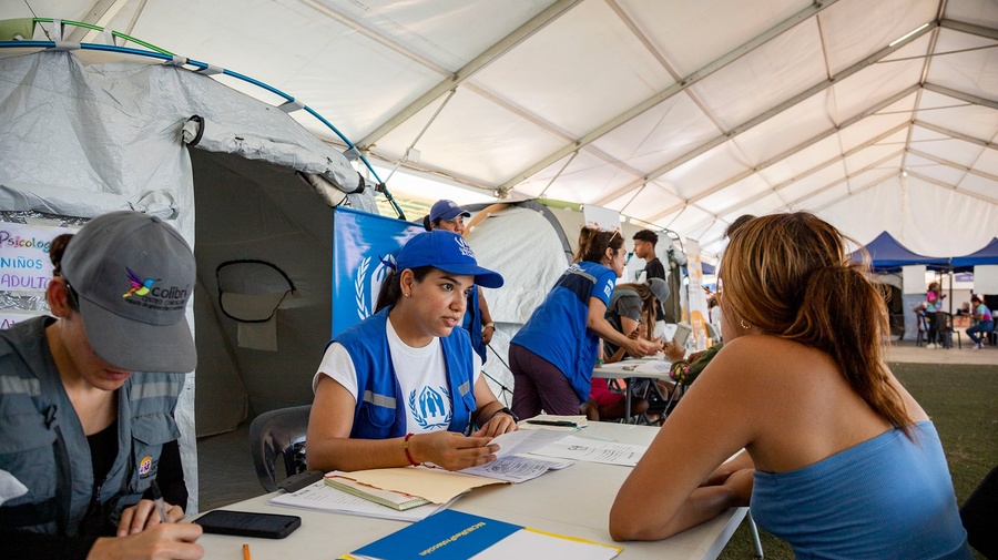 Detrás de una mesa, una trabajadora de ACNUR con vestimenta azul de la organización, atiende a una mujer que está de espaldas del otro lado de la mesa