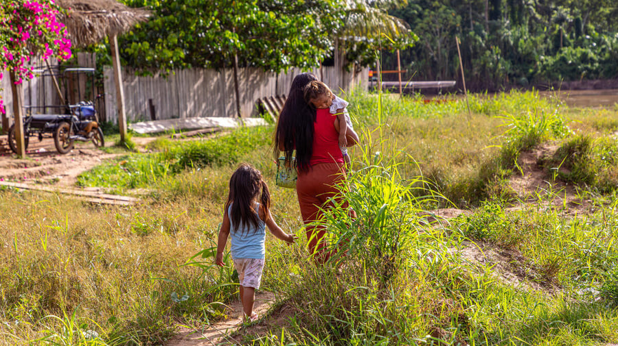 Una mujer camina de espaldas cargando en sus brazos a una niña, mientras otra niña las sigue de cerca.