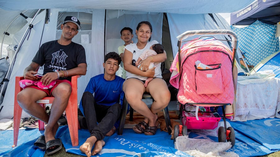 Una familia de cinco miembros, entre ellos un adolescente, un niño y una bebé, está sentada frente a la entrada de una tienda de campaña
