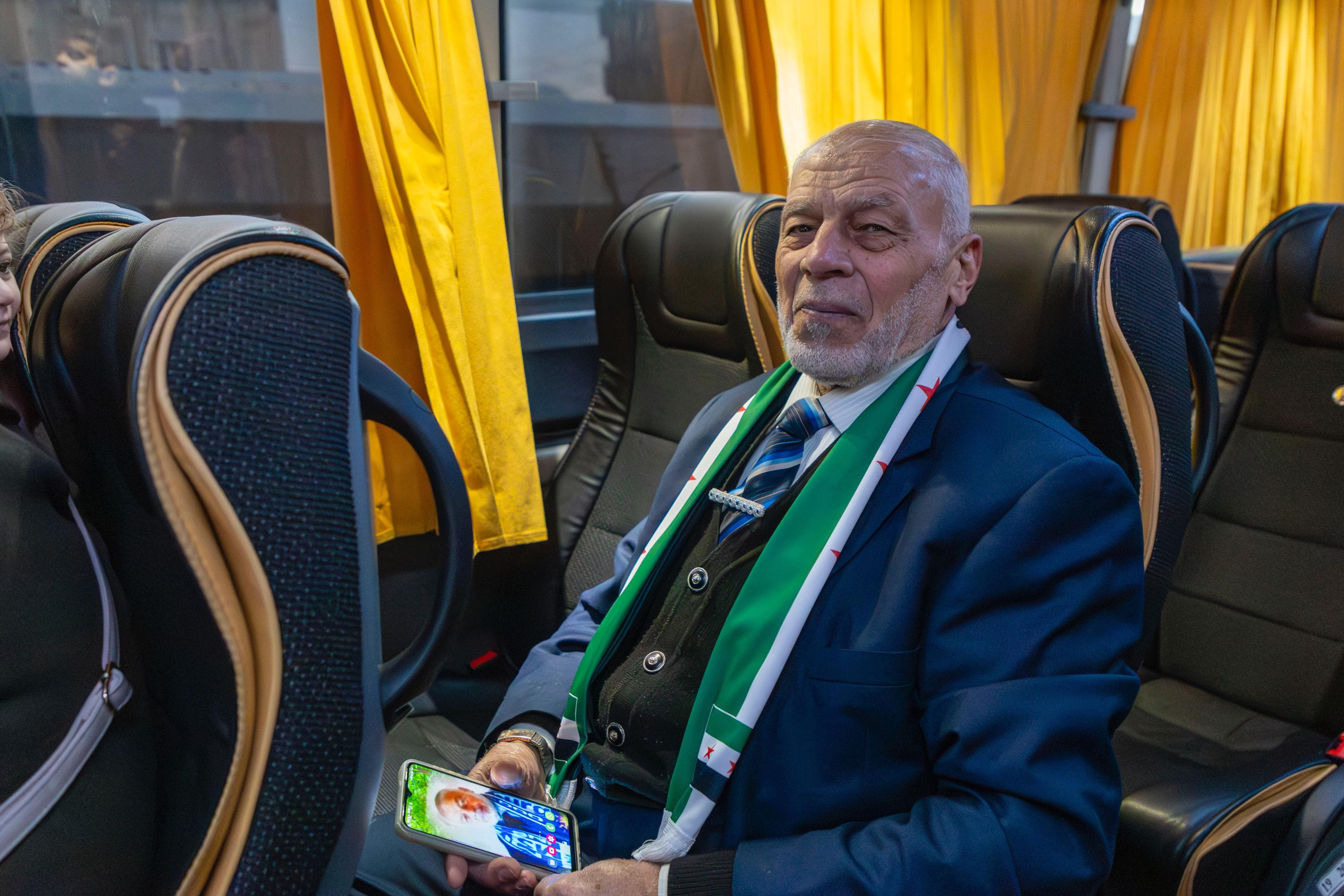 Un hombre mayor de saco y corbata sentado en un bus sujeta un celular en sus manos y sonríe mirando hacia la cámara.