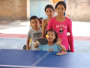 Tres niñas y un niño esperan su turno para golpear la pelota con una raqueta sobre una mesa de ping-pong color azul
