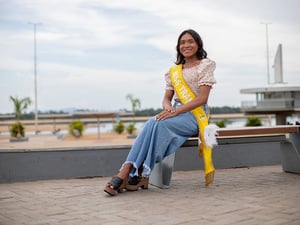 Una mujer warao, un pueblo indígena en Venezuela, sonríe a la cámara sentada en una banca en un espacio público. Lleva puesta una banda que dice "Miss Trans".