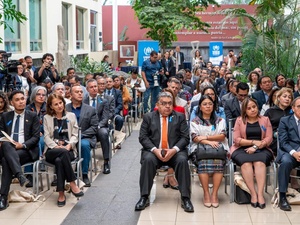 Decenas de personas sentadas en filas en un jardín. Al fondo se observan dos letreros de ACNUR en color azul