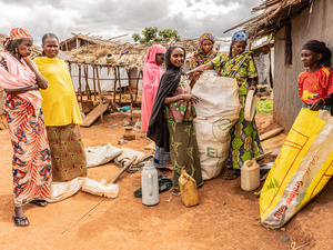 Un grupo de mujeres con vestidos de colores frente a una choza de adobe mientras llenan dos grandes sacos con sus pertenencias