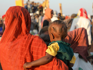 Una mujer sudanesa lleva en brazos a su bebé; se les va de espaldas. Delante suyo, un numeroso grupo de personas.
