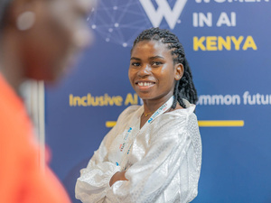 Una joven sonriente, con los brazos cruzados, posa delante del cartel de una conferencia sobre las mujeres en el ámbito de la IA en Kenia