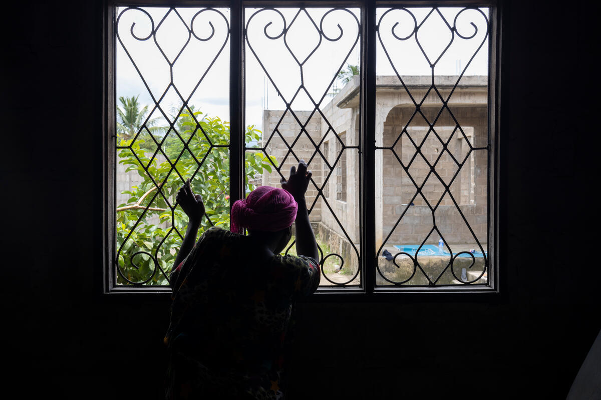 Una mujer de espaldas a la cámara mira por una ventana con las manos en los barrotes de seguridad.