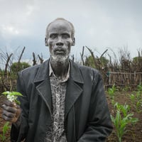 Un hombre mayor posa para una foto mostrando una planta en un campo de cultivo.