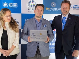 Una mujer de cabello rubio y dos hombres vestidos de traje, posan para la foto con un fondo de cuadros azules y blancos detrás