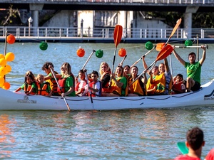 Los tripulantes de la canoa de Dársena Deportiva, decorada con guirnaldas verdes y naranjas, saludan desde el Guadalquivir.