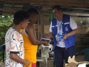 Un hombre con visibilidad de ACNUR en entrega de lámparas solares a dos hombres en una casa de madera.