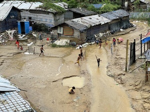 Niñas y niños juegan bajo la lluvia en un campamento de ACNUR enlodado. Se pueden ver dos alojamientos construidos con lonas y alambre