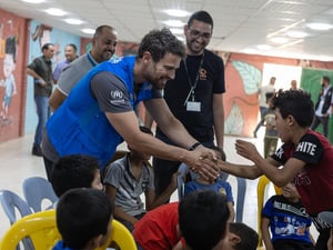 Un hombre joven que lleva puesto un chaleco con el logotipo de ACNUR salud a un grupo de niños en un campamento de refugiados en Jordania
