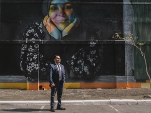 Un hombre vestido con traje y corbata de pie en una calle frente a un muro negro con el rostro de una mujer en colores
