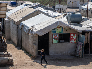 Una niña caminando delante de un alojamiento temporal de ladrillos techado con lonas de plástico en un campamento de desplazados.