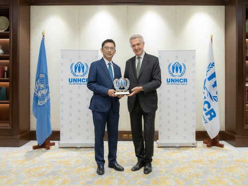 Mr. Wanchai Tachavejnukul and Filippo Grandi smiling and holding an award together, standing in front of UNHCR banners and flags