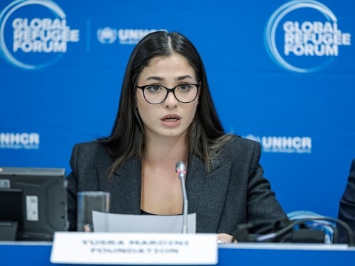 Una mujer joven, que usa anteojos, pronuncia unas palabras frente a un micrófono; detrás suyo, un fondo azul con el logotipo del Foro Mundial sobre los Refugiados.
