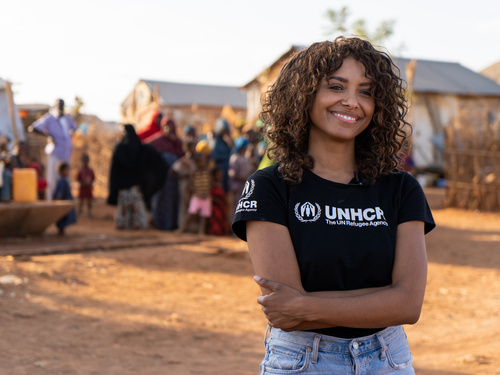 Una mujer afrodescendiente sonríe a la cámara con los brazos cruzados sobre el abdomen; detrás suyo, un grupo de personas en un campamento de refugiados