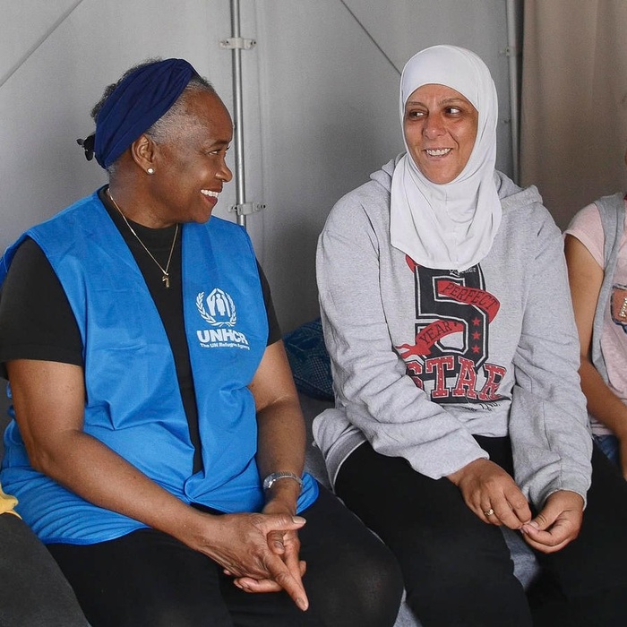 Tres mujeres sentadas una a lado de la otra se sonríen. A la izquierda, una mujer afroestadunidense que lleva puesto una chaleco azul con el logotipo de ACNUR en color blanco; a su lado derecho, una mujer que lleva puesto un jiyab color blanco.