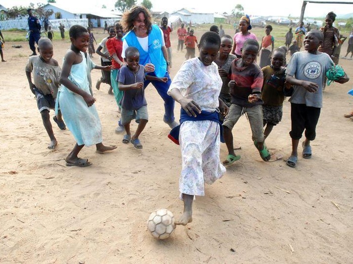Un hombre juega al fútbol con niños congoleños en un terreno polvoriento.
