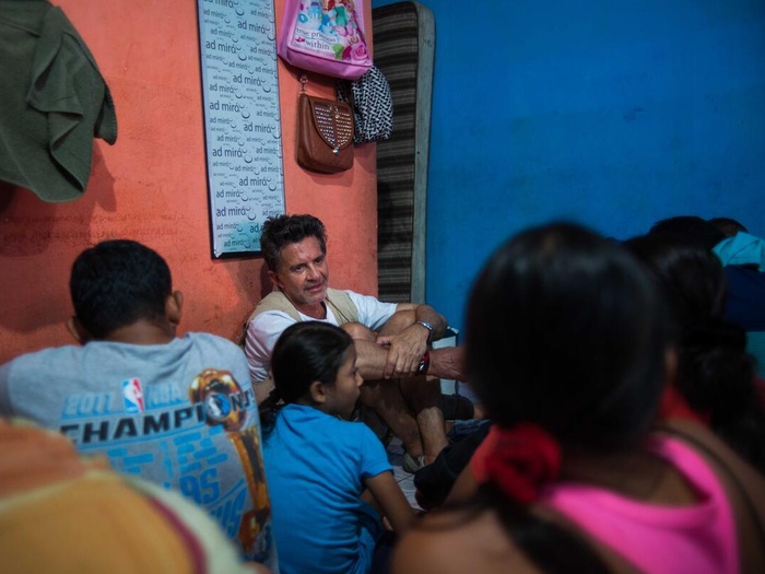 Un nombre de camiseta blanca y chaleco beige está sentado en el suelo junto a personas refugiadas y solicitantes de asilo en una habitación con paredes de colores, bolsas y un cuadro colgado detrás de él