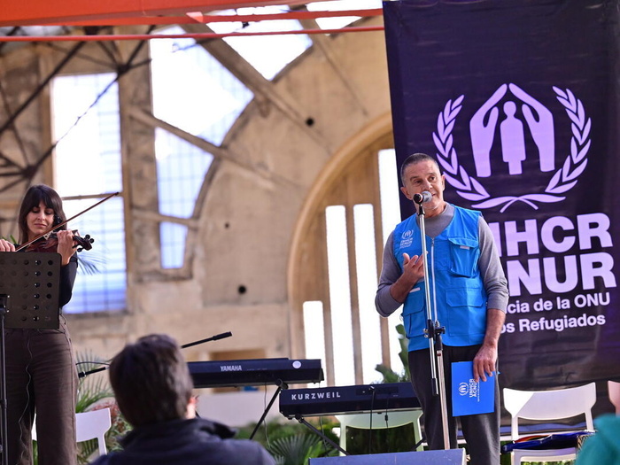 Un hombre que viste un chaleco azul de ACNUR, habla en un escenario. A sus espaldas hay una pancarta de ACNUR y a su lado un violinista, con unos teclados entre ambos