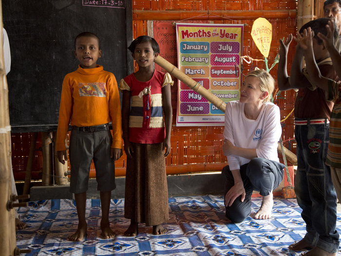 Hincada, una mujer rubia observa con cariño a un chico y una chica rohingya