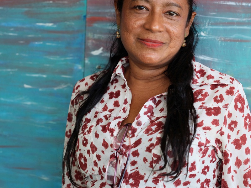 Una mujer de tez morena y cabello largo negro, que viste una blusa blanca con flores rojas, mira hacia la cámara. Al fondo, una pintura naranja con azul