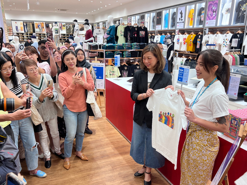 En una tienda de ropa, frente al mostrador, dos mujeres sostienen una camiseta con un estampado; frente a ellas, una multitud que toma fotografías de la prenda.