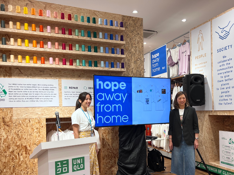 Dos mujeres junto a una pantalla que muestra una imagen de las camisetas estampadas y la leyenda "Esperanza lejos del hogar", que es el lema del concurso de arte