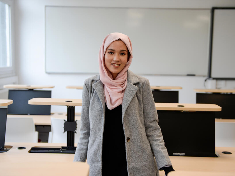 Una joven de Afganistán sonríe a la cámara desde el centro de un aula. Lleva puesto un hijab color durazno; detrás suyo, un pintarrón.