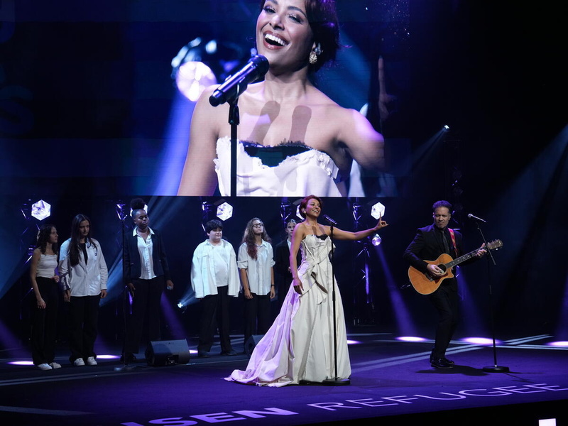 Una mujer en el escenario con un guitarrista y un coro juvenil delante de una pantalla gigante