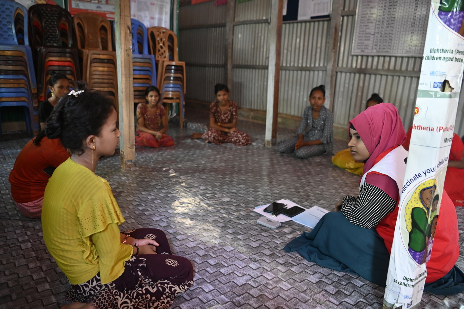 Una joven voluntaria que porta un pañuelo rosa en la cabeza, dirige una sesión de apoyo entre iguales para niñas y niños refugiados rohingyas que se encuentran sentados en el suelo en círculo