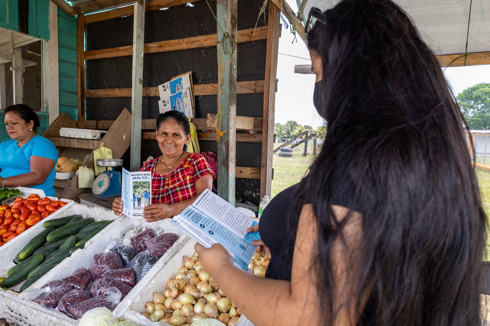 Una joven de cabello largo y oscuro entrega folletos a dos mujeres en una tienda de verduras