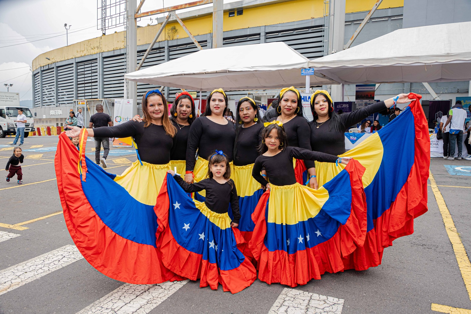 Seis mujeres y dos niñas vestidas con blusa negra y falda en colores amarillo, azul y rojo, posan para la foto afuera de un estadio