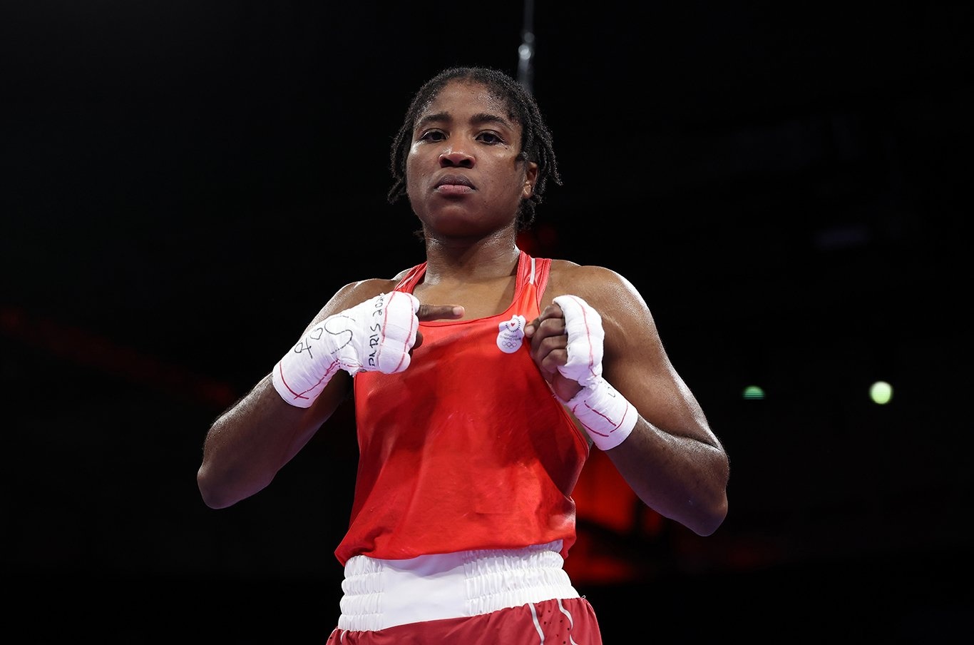 Una mujer posa para una foto con ropa de boxeo color rojo, señalando el logotipo del Equipo Olímpico de Atletas efugiados en su ropa.