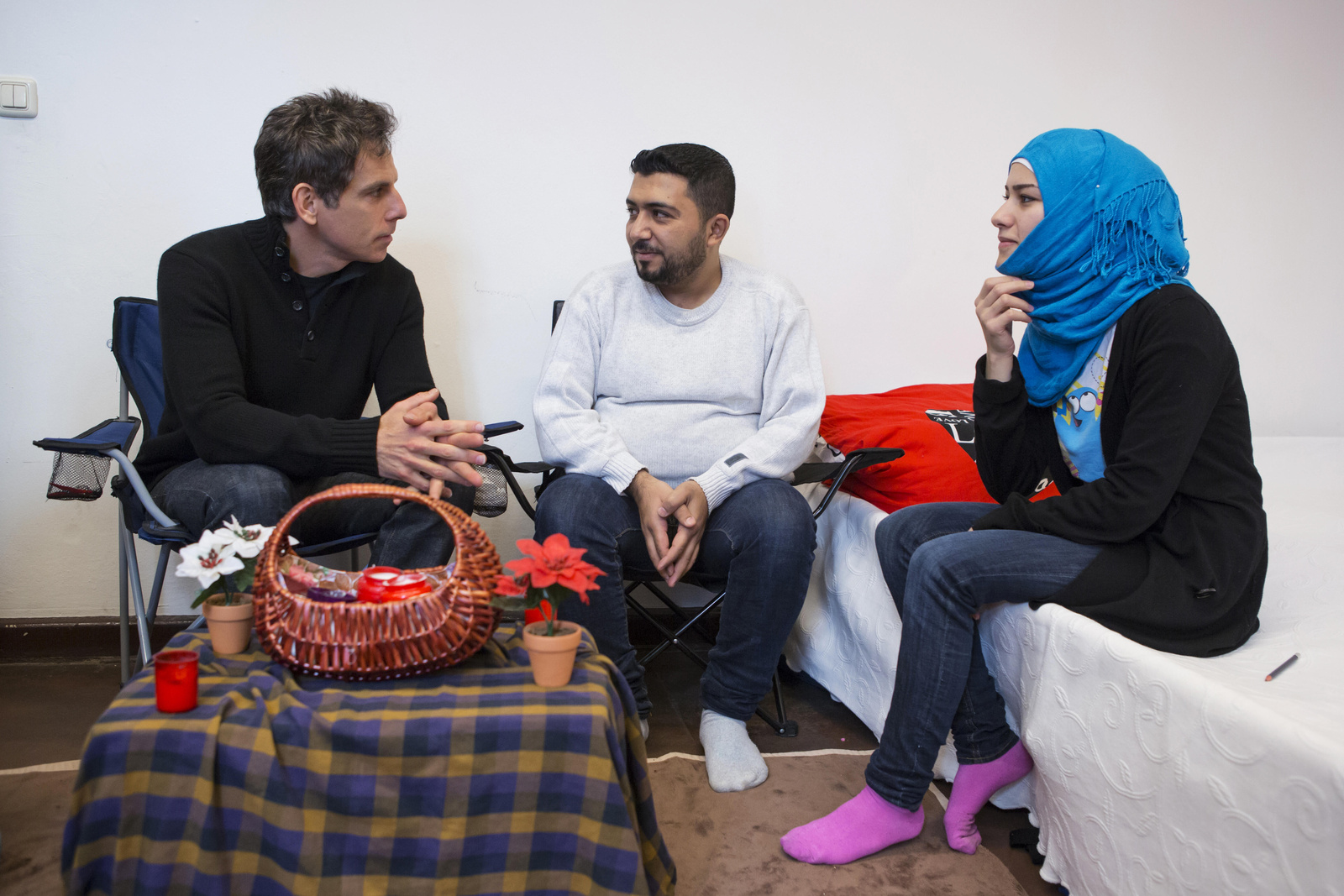 Un hombre caucásico charla con un hombre y una mujer de Siria. Ella está sentada sobre una cama y lleva puesto un hiyab.