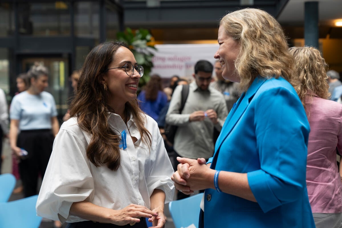 Dos mujeres, una de cabello negro con blusa blanca, y otra de cabello rubio que viste de azul, conversan sonrientes