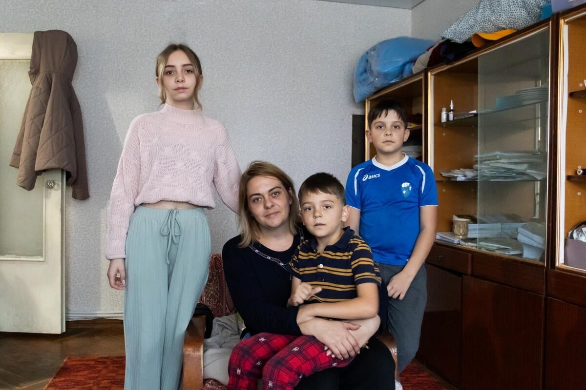 Una mujer posa para una foto con sus tres hijos en su casa. El más pequeño está sentado en su regazo, mientras que los otros dos están de pie junto a ella