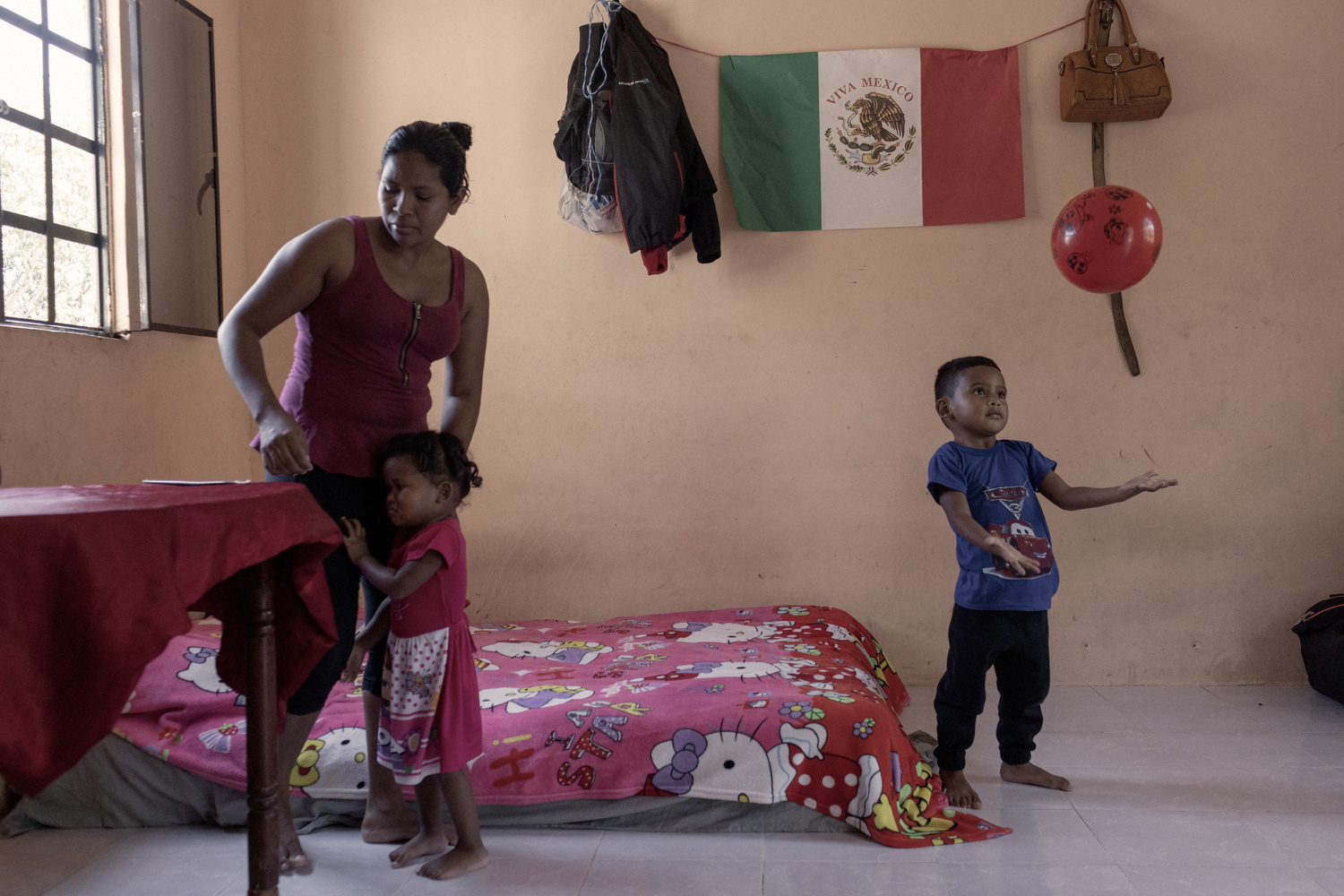 Una mujer atiende a una niña pequeña mientras un niño juega con una pelota roja.