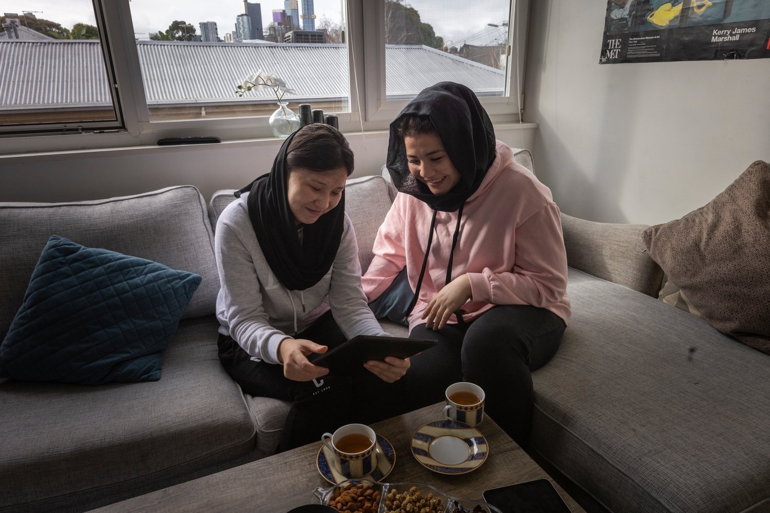 Dos jóvenes están sentadas en un sillón, una junto a la otra, observan un tableta y toman té.