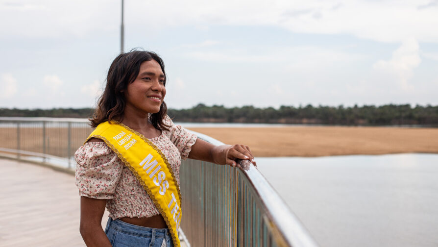 Una mujer warao, un pueblo indígena en Venezuela, sonríe viendo al horizonte. Está recargada en un barandal y lleva puesta una banda que dice "Miss Trans".