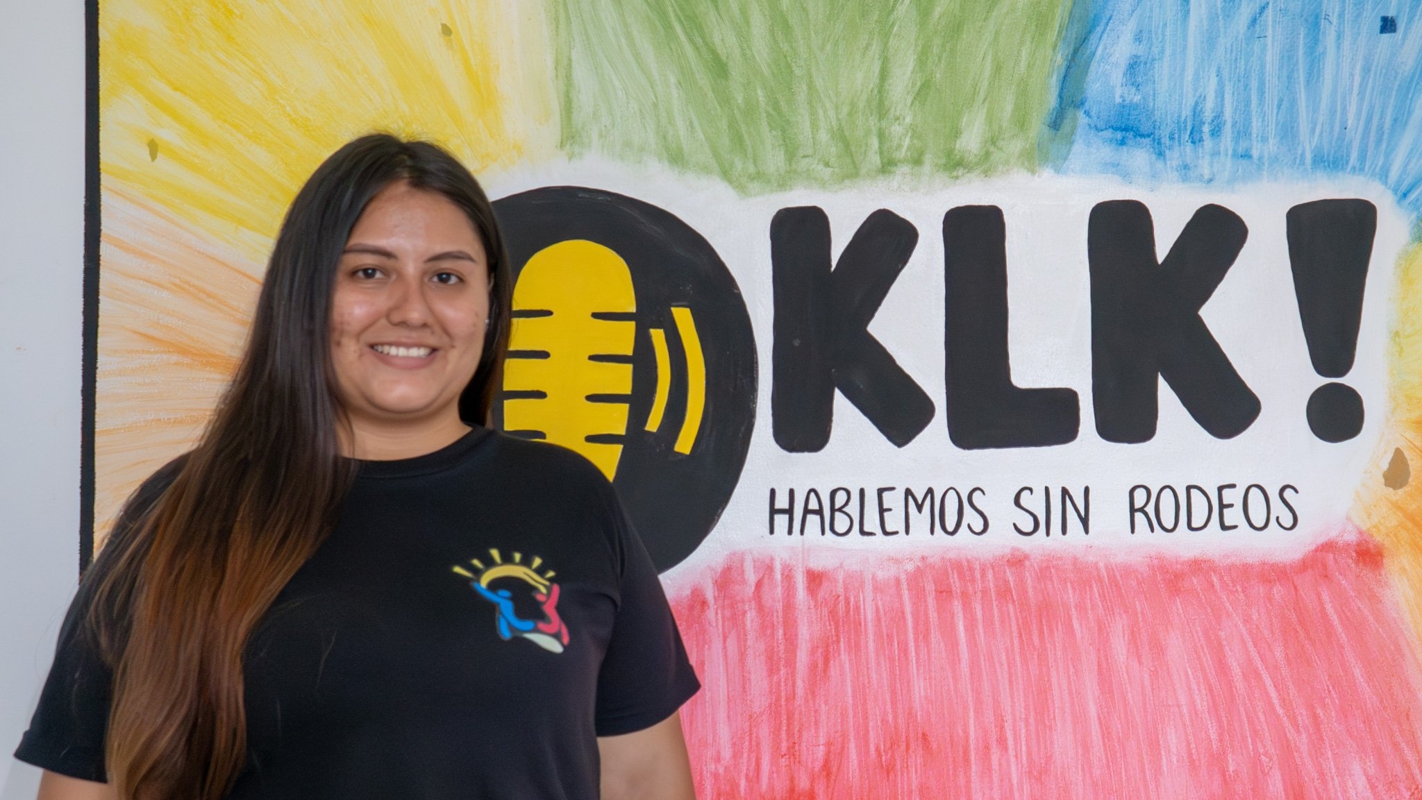 Una joven posa sonriendo para una foto delate de un mural donde se lee &iexcl;KLK, hablemos sin rodeos!