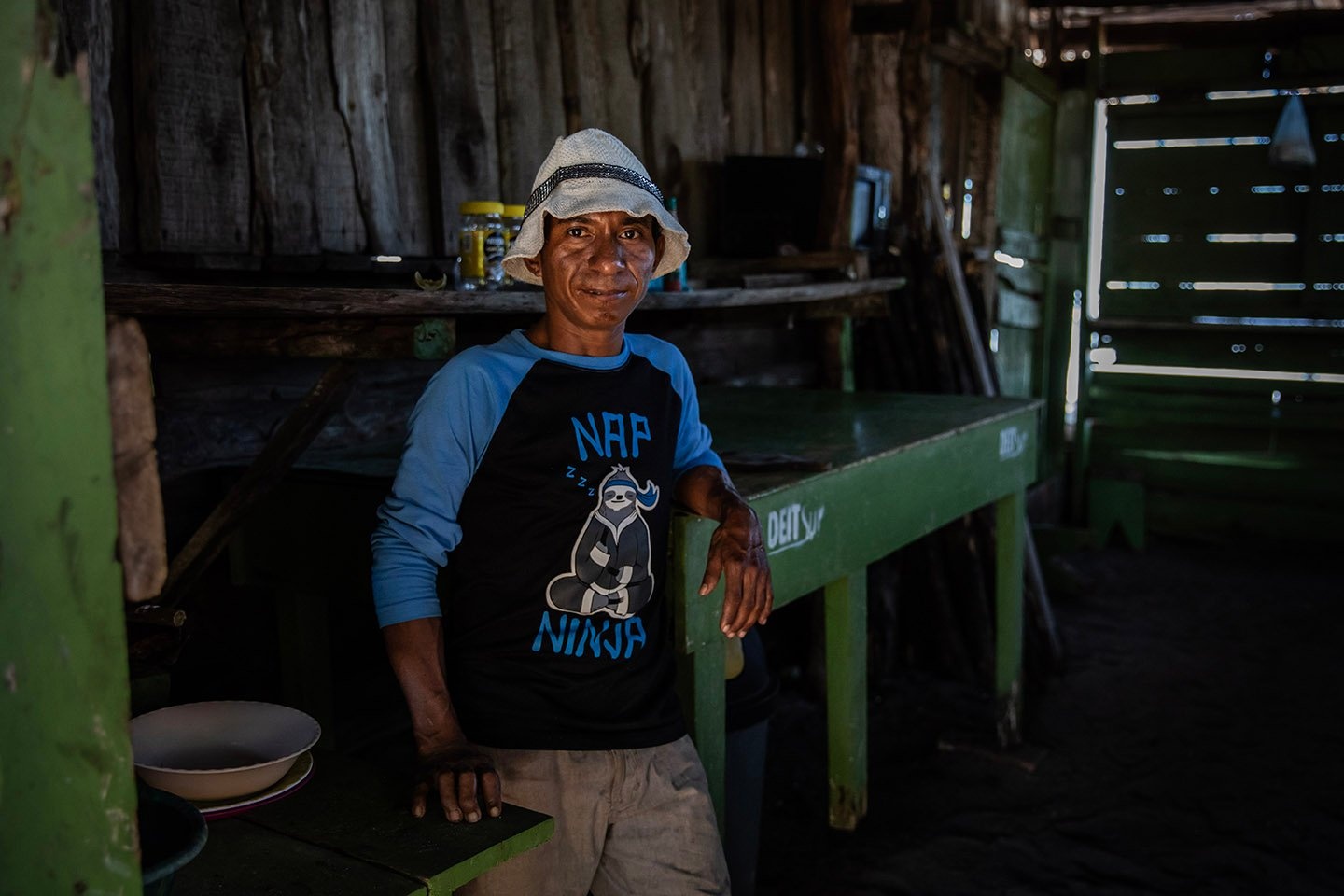 Un hombre con un sombrero de pescador posa frente a cámara en una sencilla casa de madera.